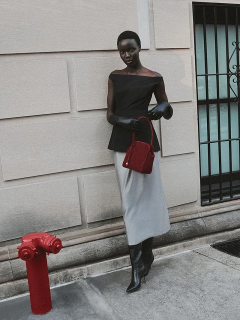 Elegant woman in a black top, gray skirt, and black boots stands next to a red fire hydrant on a city sidewalk with neutral wall.