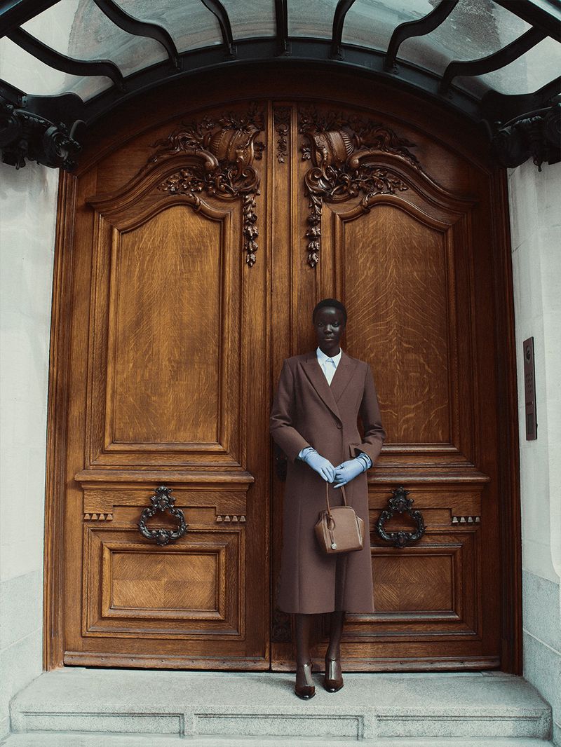 Woman in brown coat and blue gloves poses in front of large wooden doors with intricate carvings and a wrought iron transom.