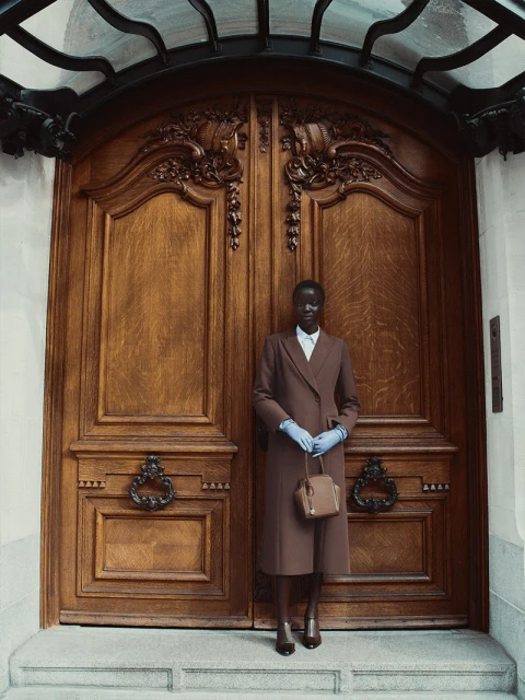 Woman in brown coat and blue gloves poses in front of large wooden doors with intricate carvings and a wrought iron transom.