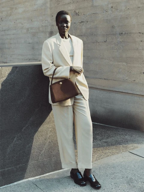 Elegant dark-skinned woman in cream pantsuit with brown handbag stands against a textured wall in a fashionable outdoor setting.