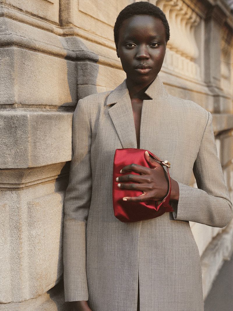 Portrait of an African female model wearing a grey blazer and holding a red satin clutch bag against a stone wall.