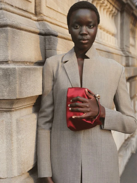 Portrait of an African female model wearing a grey blazer and holding a red satin clutch bag against a stone wall.