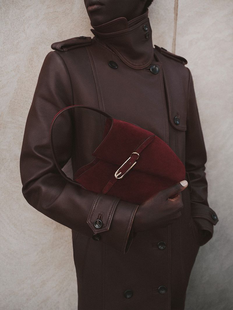 A person with dark skin wearing a leather trench coat holding a burgundy velvet purse with a gold clasp against a neutral backdrop.