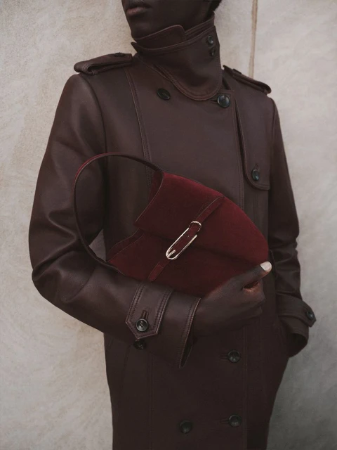 A person with dark skin wearing a leather trench coat holding a burgundy velvet purse with a gold clasp against a neutral backdrop.