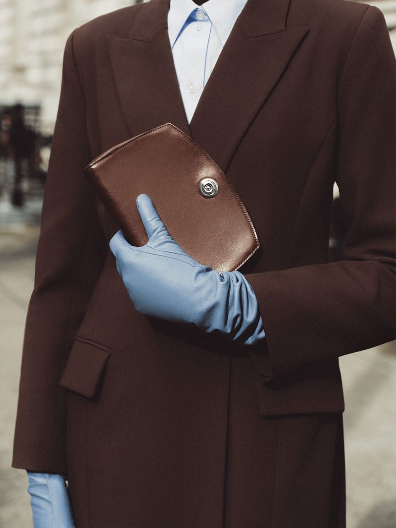Fashion shot of a woman in a brown suit and blue gloves holding a brown clutch against a neutral urban background.