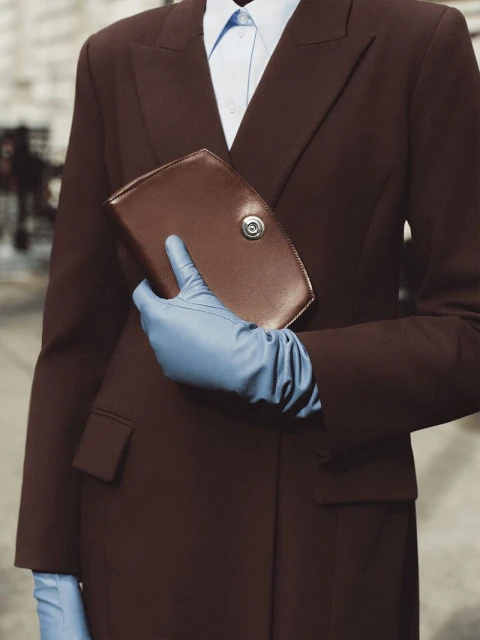 Fashion shot of a woman in a brown suit and blue gloves holding a brown clutch against a neutral urban background.