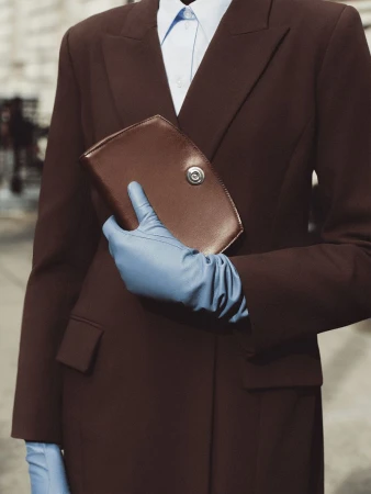 Fashion shot of a woman in a brown suit and blue gloves holding a brown clutch against a neutral urban background.