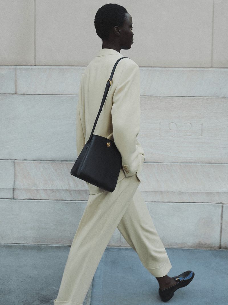 Fashionable woman in light suit walks with black leather satchel bag, pairing it with black loafers on a sidewalk beside a wall.