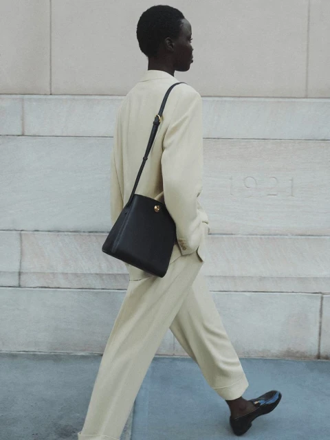 Fashionable woman in light suit walks with black leather satchel bag, pairing it with black loafers on a sidewalk beside a wall.