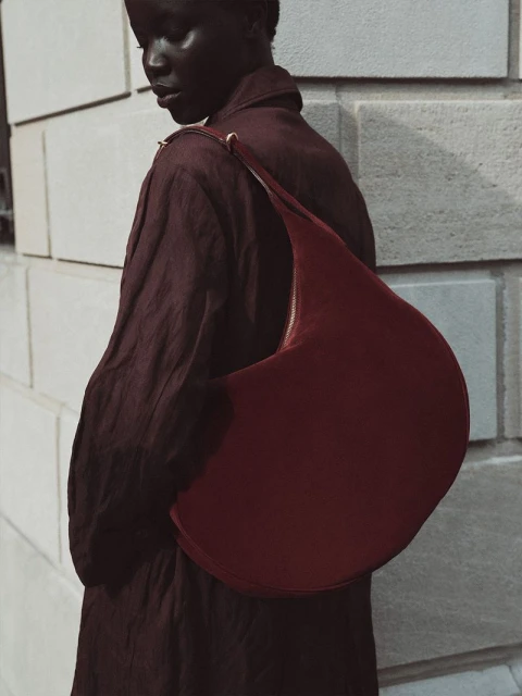Woman in burgundy outfit carrying large burgundy crescent bag standing against a textured light-grey wall in soft natural light.