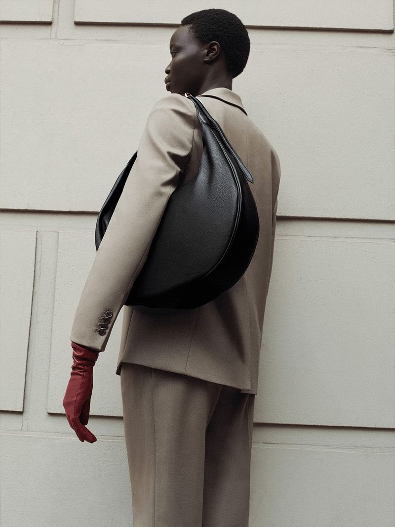 Model wearing a tailored beige suit and red leather gloves carrying a large black curved leather shoulder bag against a paneled wall.