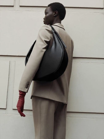 Model wearing a tailored beige suit and red leather gloves carrying a large black curved leather shoulder bag against a paneled wall.
