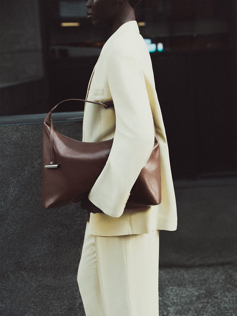 Model wearing a tailored cream suit carrying a slouchy brown leather shoulder bag against an urban backdrop.