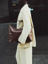 Model wearing a tailored cream suit carrying a slouchy brown leather shoulder bag against an urban backdrop.