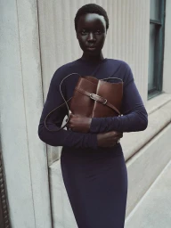 Black woman with short buzzcut hair wearing a navy blue long-sleeve dress and holding a brown leather bucket bag against a stone wall.