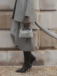 Cropped shot of a model in a grey tailored skirt suit and black stiletto boots holding a small animal print handbag.