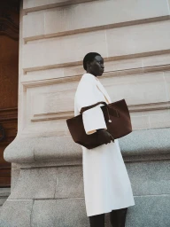 Model in a white coat holding a large brown tote bag standing against a classical stone building exterior