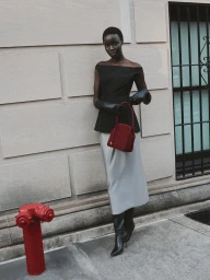 Model in a black off-the-shoulder top and grey satin skirt holding a burgundy handbag on a sidewalk beside a red fire hydrant.