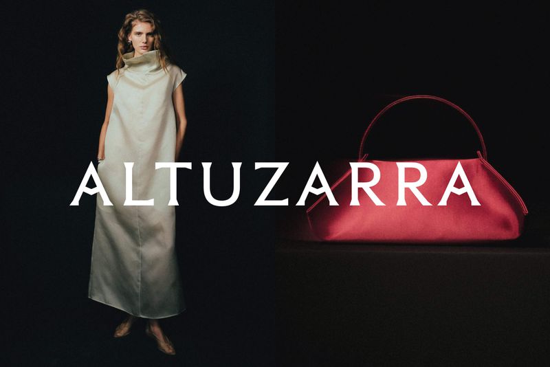 Fashion shot of a woman in a long white dress standing next to a red handbag against a dark background, featuring the brand name Altuzarra.