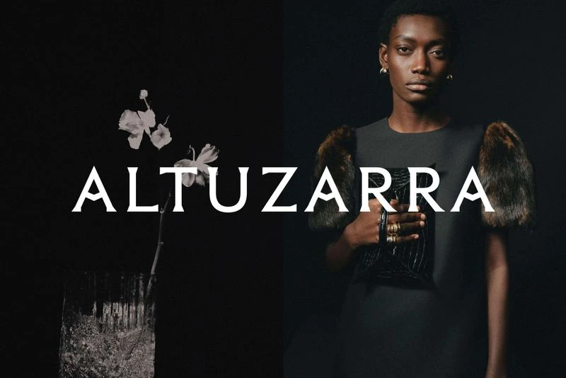 Fashion shot of a Black woman in dress with fur sleeves, holding a black purse, against a dark backdrop. 'Altuzarra' text overlay.