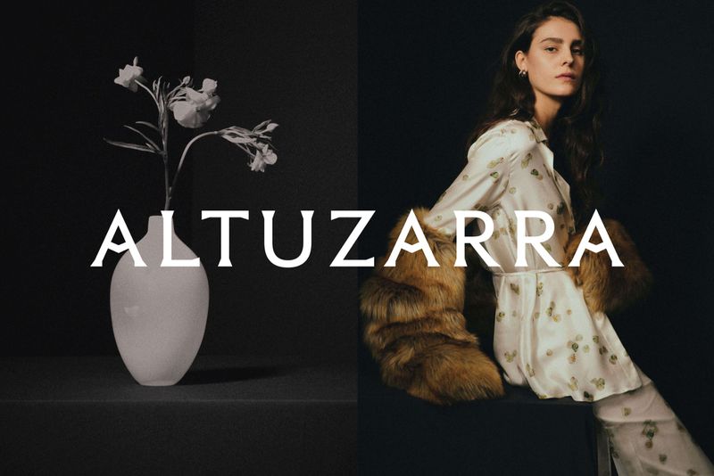 Model in a patterned white silk shirt and brown fur stole seated beside a white vase with flowers under the bold typography ALTUZARRA.