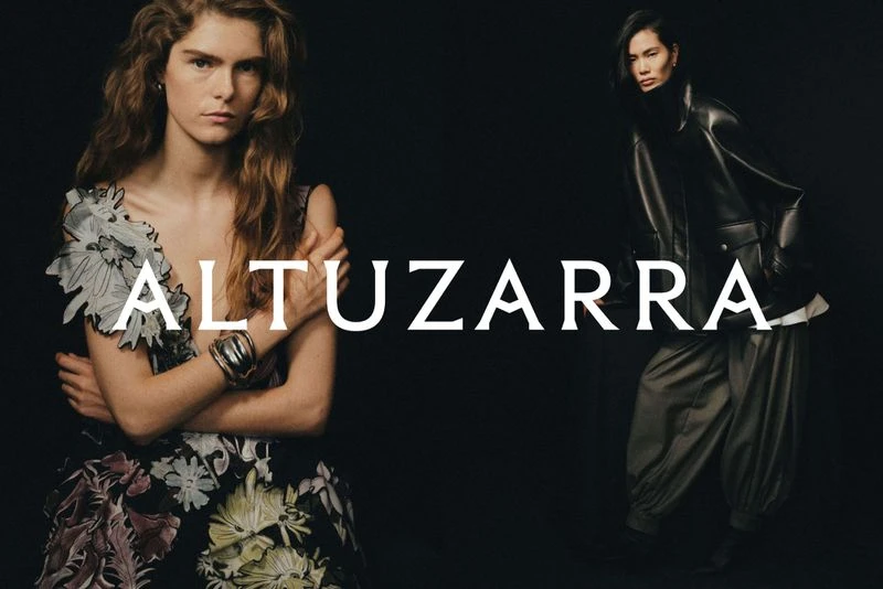 Two women in a fashion editorial shot against a black background. One woman wears a floral dress, and the other wears a black leather jacket.
