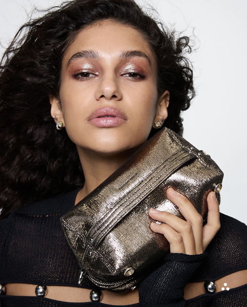 Glamorous woman poses with a metallic clutch, showcasing bold eyeshadow and elegant earrings against a stark white background.