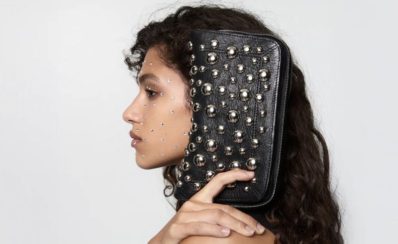 Profile of a woman with dark curly hair showcasing a black leather clutch purse adorned with silver studs against a clean white background.