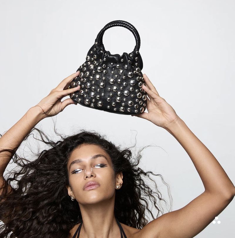 Model poses with a black leather studded bucket bag against a white backdrop, showcasing fashion and accessorizing.