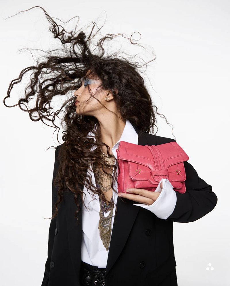 Tamar David holding a vibrant pink leather handbag with windblown curly hair wearing a black blazer against a white backdrop.