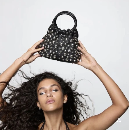 Model Tamar David holding a black studded leather handbag above her head against a plain white studio background.