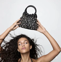 Model Tamar David holding a black studded leather handbag above her head against a plain white studio background.