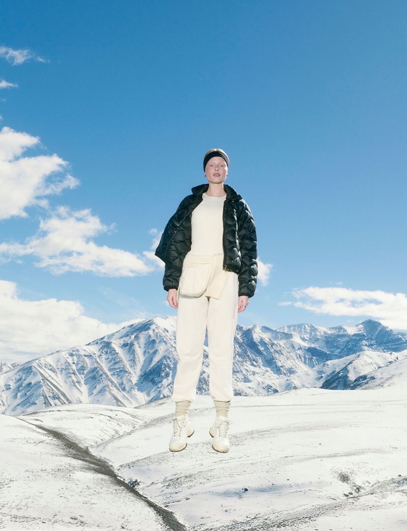 Female model posing in a cream outfit and puffer jacket against a bright sky and snowy mountain backdrop on a sunny day.