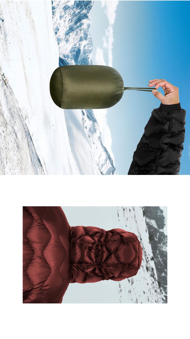 Person holding a packed olive-green sleeping bag in a snowy mountain landscape under a blue sky with white clouds. Quilted jacket visible.