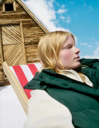 Woman relaxing in a red and white striped deckchair on a snow-covered surface near a rustic wooden cabin against a blue sky.