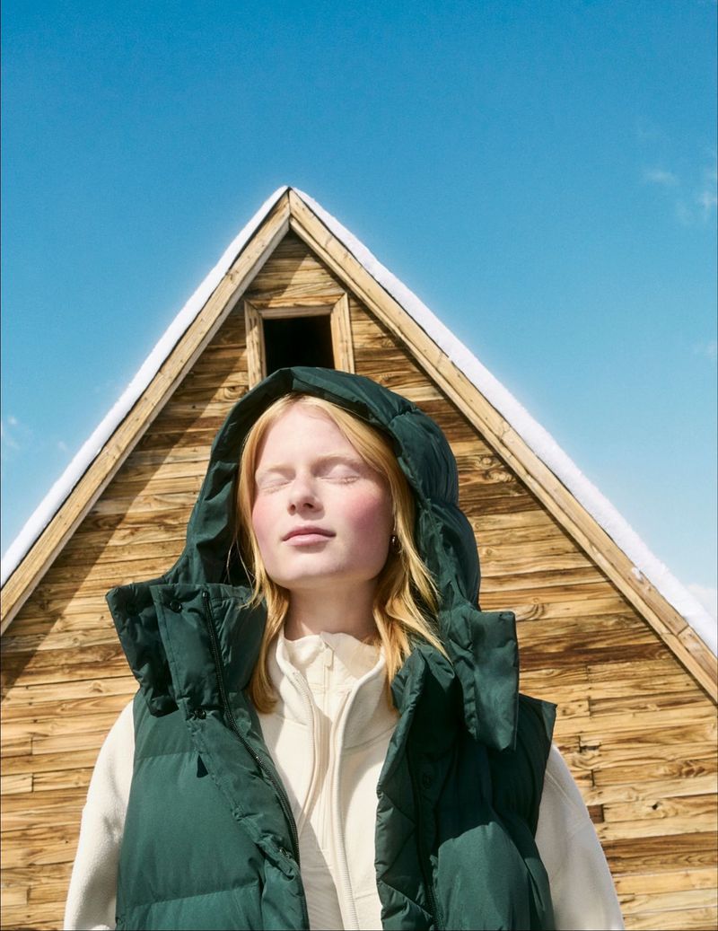 Young woman with blonde hair and closed eyes wearing a hooded puffer vest outdoors with a wood cabin and blue sky in the background.
