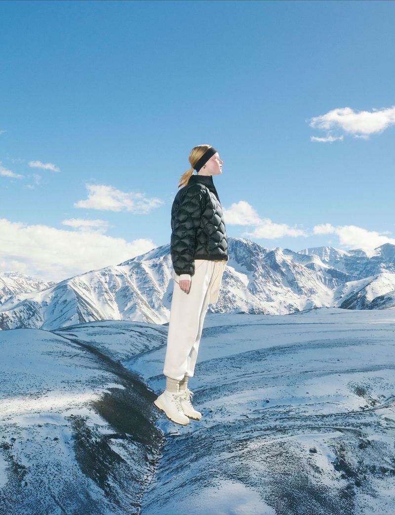 Woman in black puffer jacket and white pants in a snowy mountain landscape under a blue sky with scattered clouds. Fashion shot.