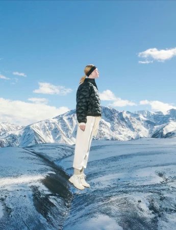 Woman in black puffer jacket and white pants in a snowy mountain landscape under a blue sky with scattered clouds. Fashion shot.