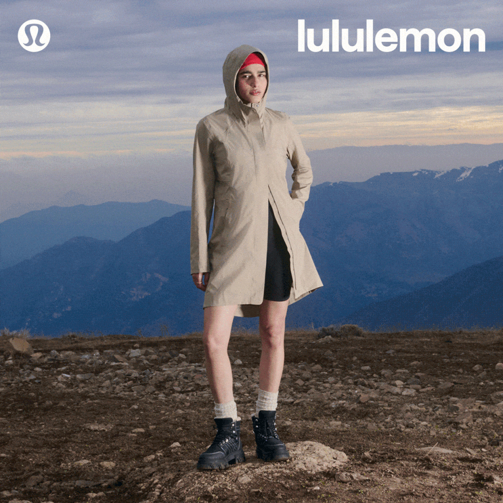 A woman in a beige raincoat standing on a rocky outcrop with snow-capped mountains and cloudy skies in the background, wearing Lululemon.