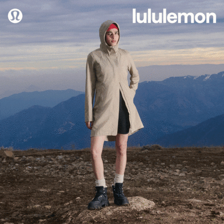A woman in a beige raincoat standing on a rocky outcrop with snow-capped mountains and cloudy skies in the background, wearing Lululemon.
