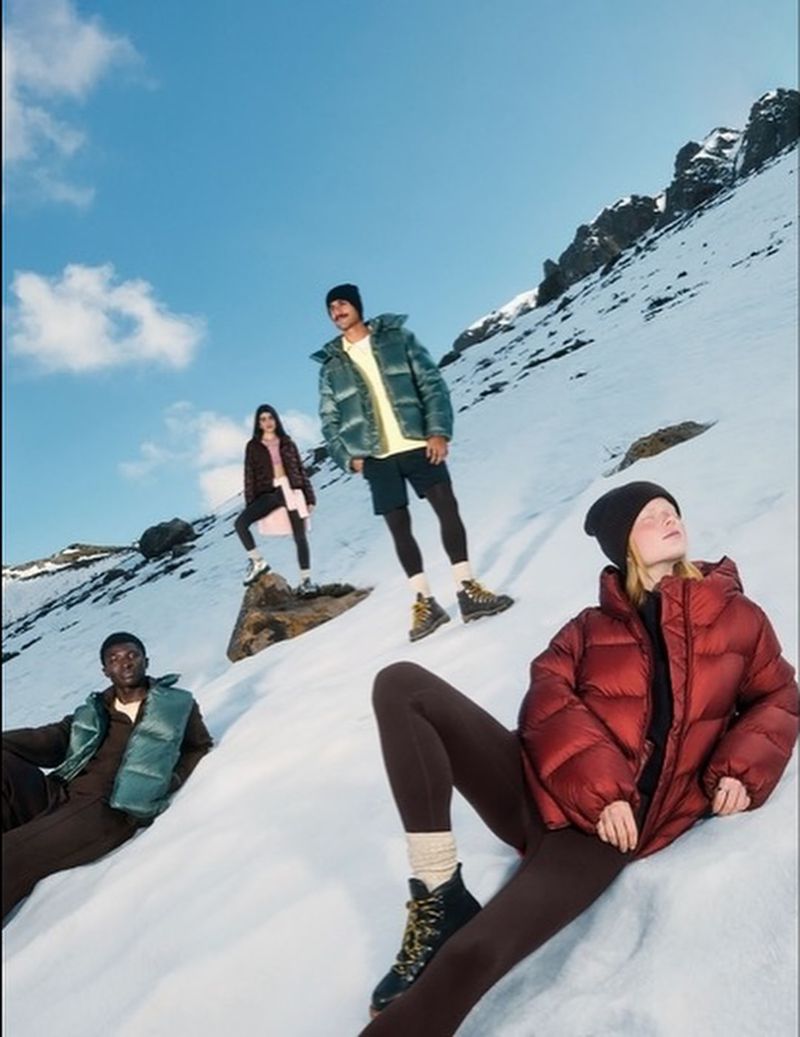 Group of four diverse individuals in puffy jackets pose in snowy mountain landscape on a sunny day, fashion editorial photography.