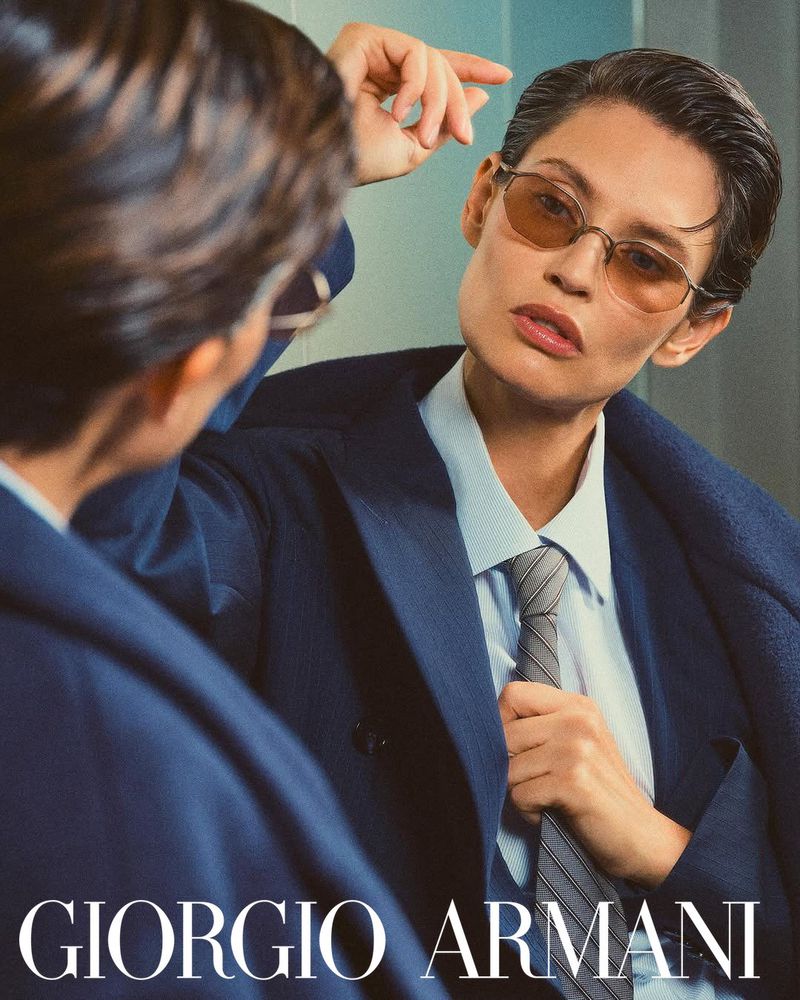 Fashion shot of a woman in a blue suit and tan glasses, reflected in a mirror, with the Giorgio Armani logo visible at the bottom.