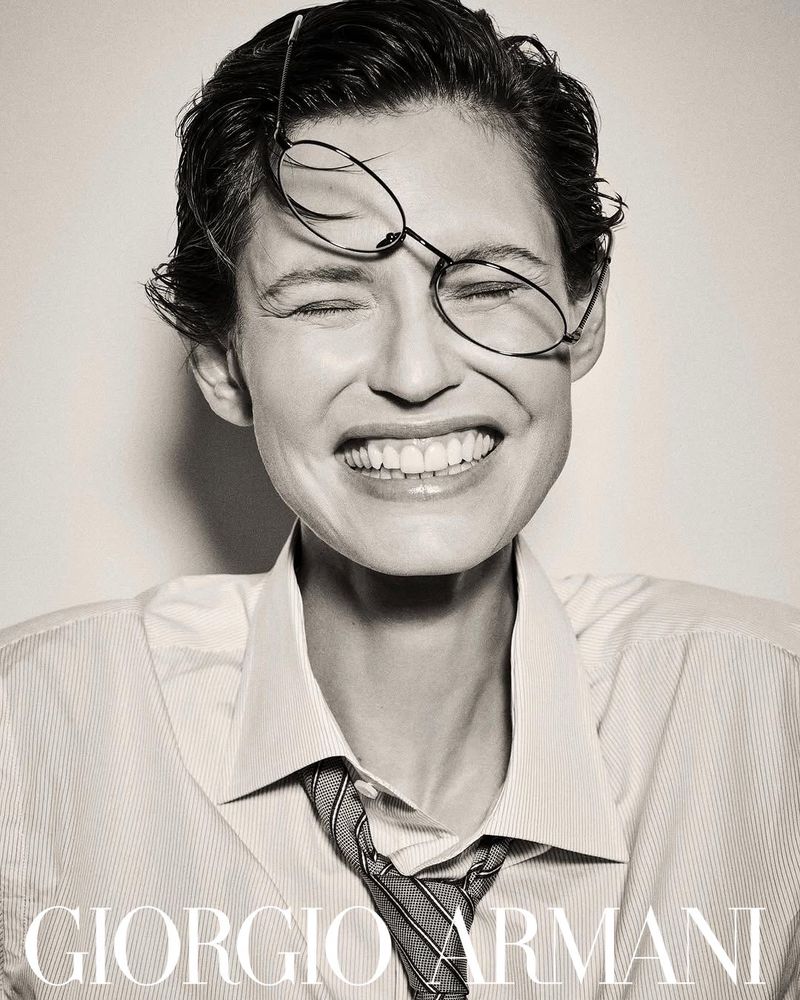 Close-up of a smiling woman with glasses on her head, wearing a striped shirt and patterned tie, featuring the Giorgio Armani logo.