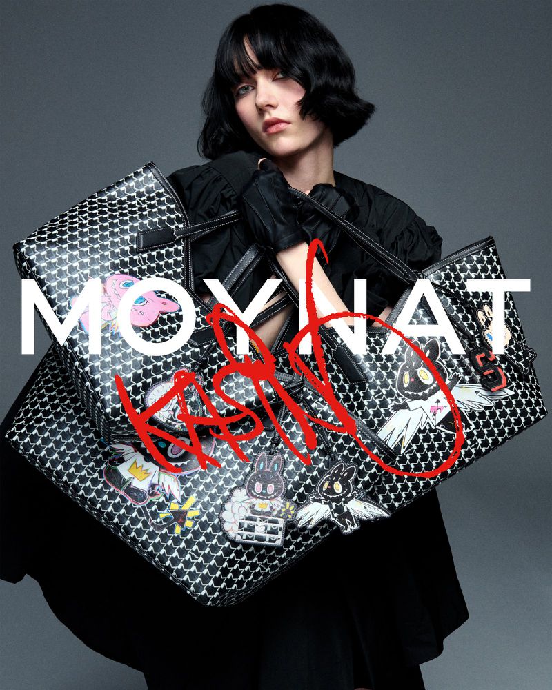 Fashion shot of a female model with bob haircut holding two printed Moynat tote bags against a grey backdrop in a studio.