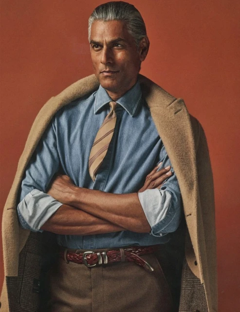 Portrait of adult man wearing a blue button-down shirt, brown pants, tie, and jacket, arms crossed against a solid orange backdrop.