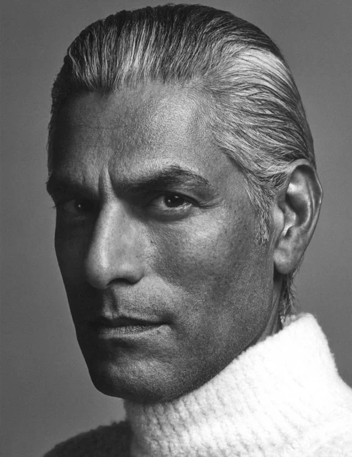 Close-up portrait of a tanned, gray-haired adult male wearing a white turtleneck sweater against a neutral gray background.