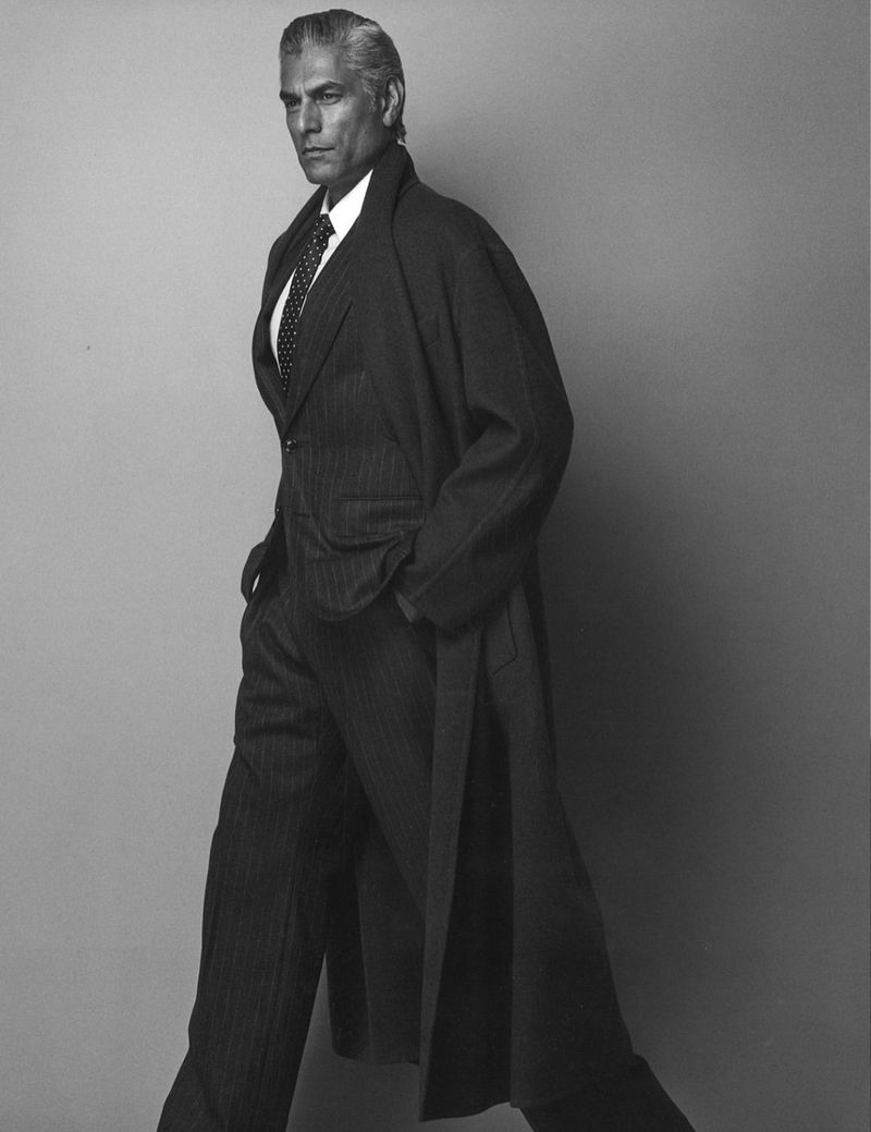 Black and white studio portrait of an adult male in a striped suit with a tie, wearing a long dark overcoat, walking and looking at camera.