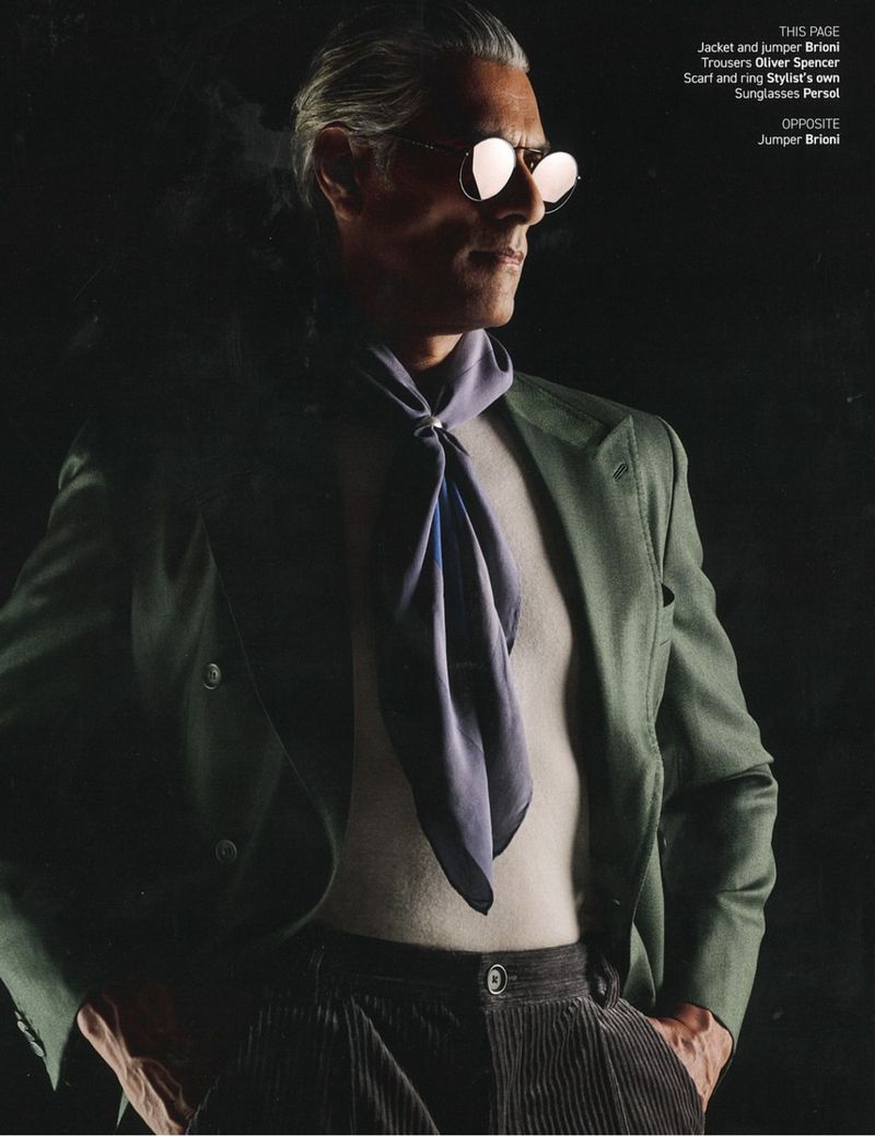 Middle-aged man poses in stylish olive jacket, light sweater, corduroy trousers, and aviator sunglasses against a dark background.