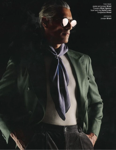 Middle-aged man poses in stylish olive jacket, light sweater, corduroy trousers, and aviator sunglasses against a dark background.