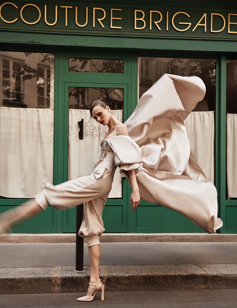 Fashion editorial: Woman poses in an elegant beige outfit with flowing fabric in front of a green building with gold lettering outdoors.